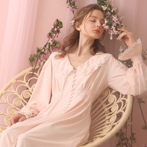Beautiful seated woman in long, soft Margaret Lawton Nightgown. Cloth covered buttons down the front of very feminine pink nightgown with long cuffed poet sleeves and turned down collar trimmed in lace. Elegant. 